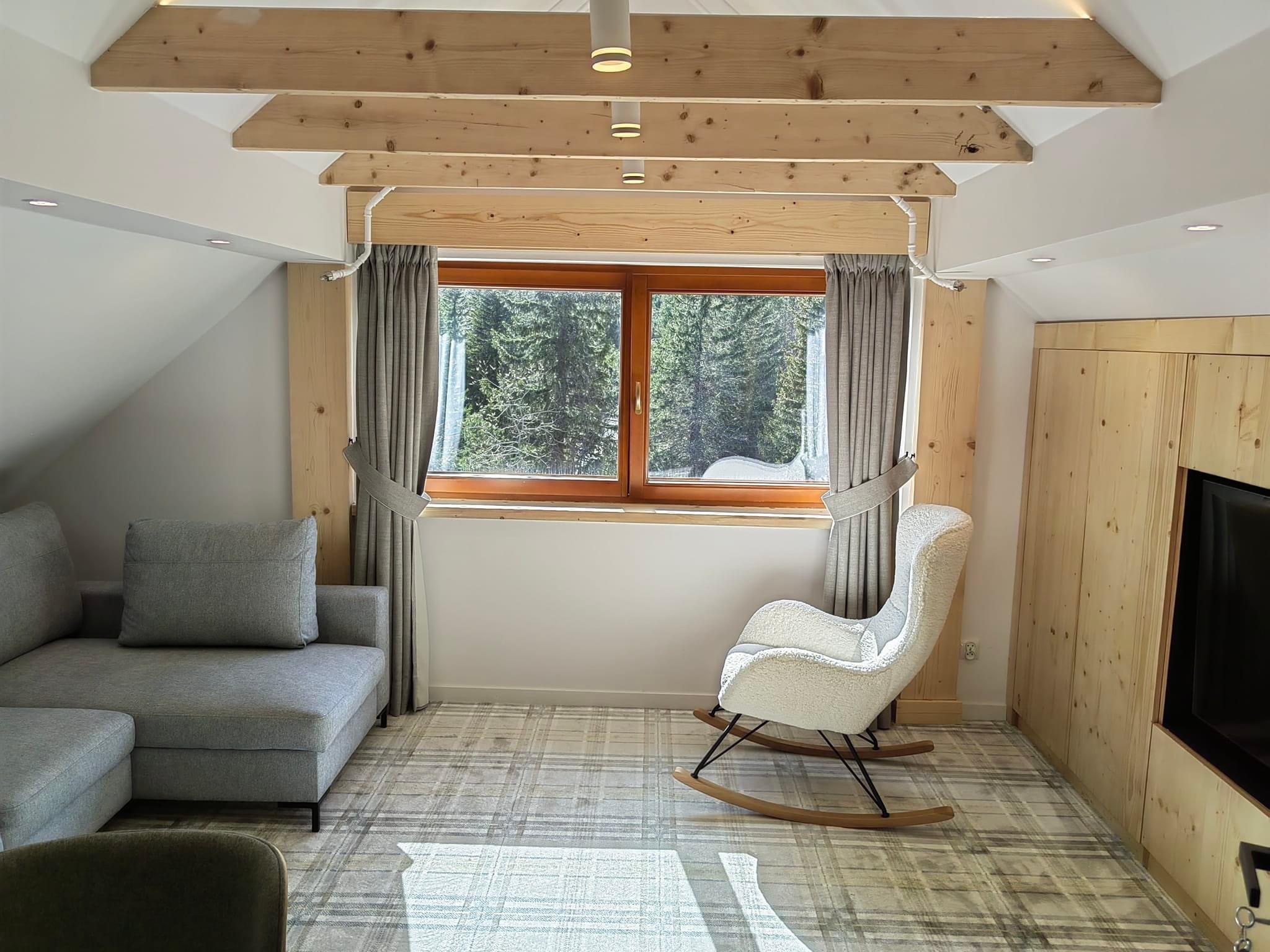 Jasne poddasze z drewnianymi belkami stropowymi, szarą sofą narożną, bujanym fotelem typu baranek i widokiem na las za oknem.