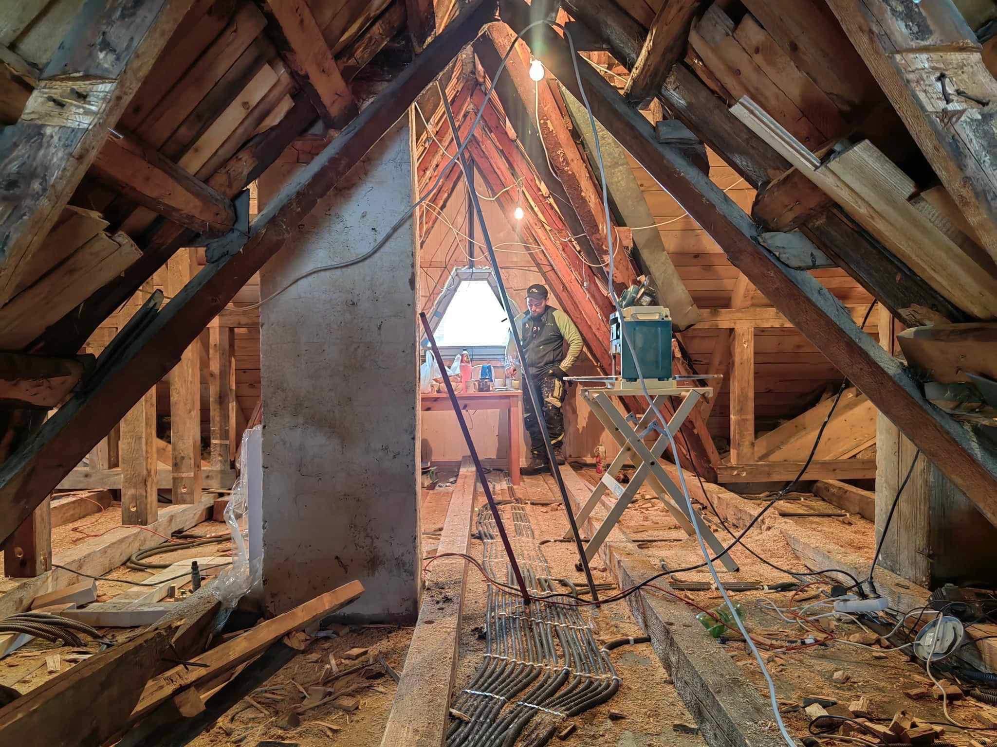 Poddasze w trakcie prac remontowych, widoczne drewniane krokwie, fragment ściany, narzędzia i przewody elektryczne, majster w tle przy oknie.