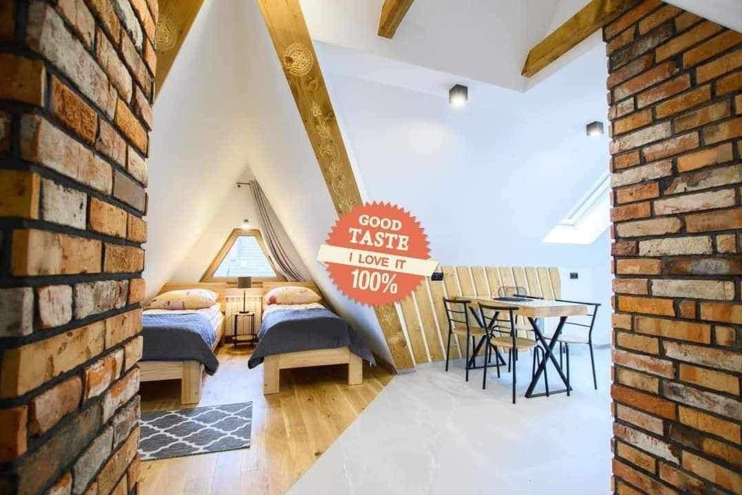 Pokój na poddaszu z dwiema pojedynczymi łóżkami, drewnianą podłogą, małym oknem w kształcie trójkąta, ceglanymi filarami i stołem z krzesłami, całość w jasnej kolorystyce z naklejką 'Good Taste...