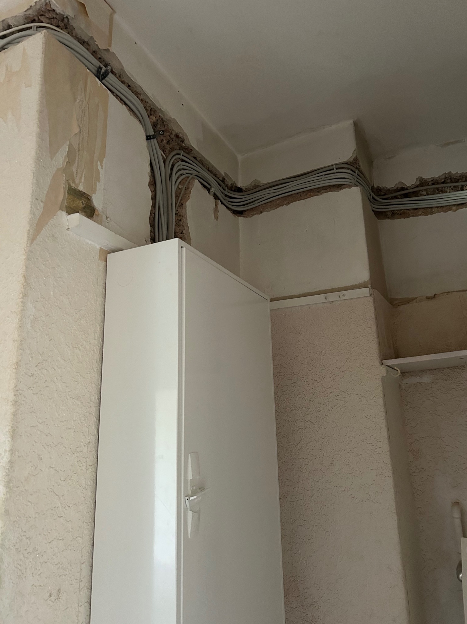 Szereg szarych kabli elektrycznych prowadzonych wzdłuż ściany pod sufitem, zabezpieczonych czarnymi uchwytami, nad białą szafką w pomieszczeniu w trakcie remontu.