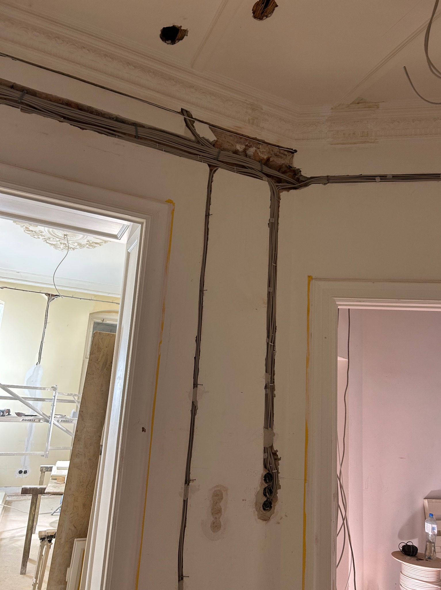 Kable elektryczne poprowadzone po ścianie w starym budynku, widoczne puszki i elementy dekoracyjne sufitu. Prace w trakcie remontu, widoczne zabezpieczenia na futrynach.