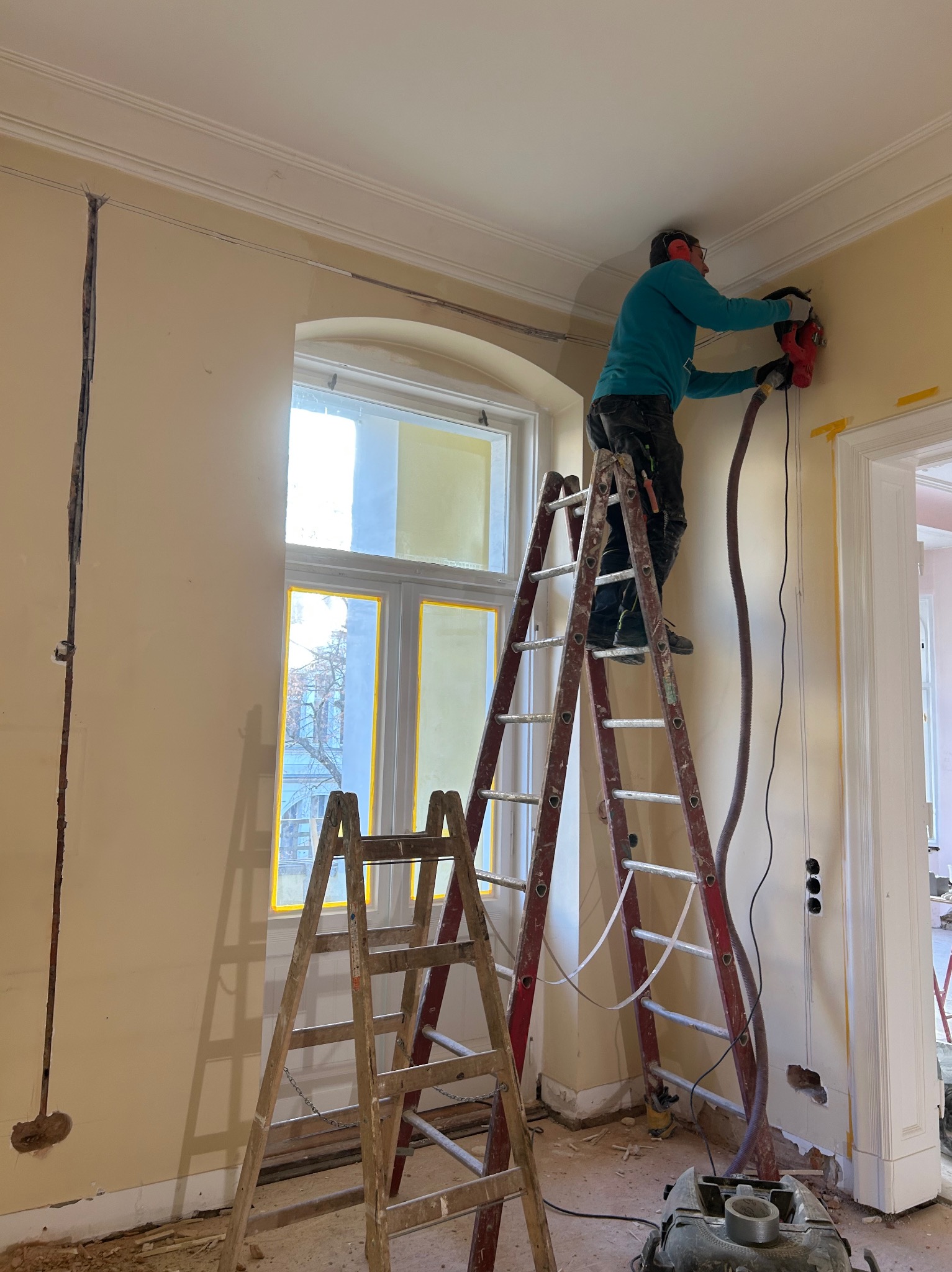 Pracownik na drabinie bruzduje ścianę pod instalację elektryczną. Widoczne kable i otwory w ścianie, odkurzacz przemysłowy. Praca w trakcie wykonywania.