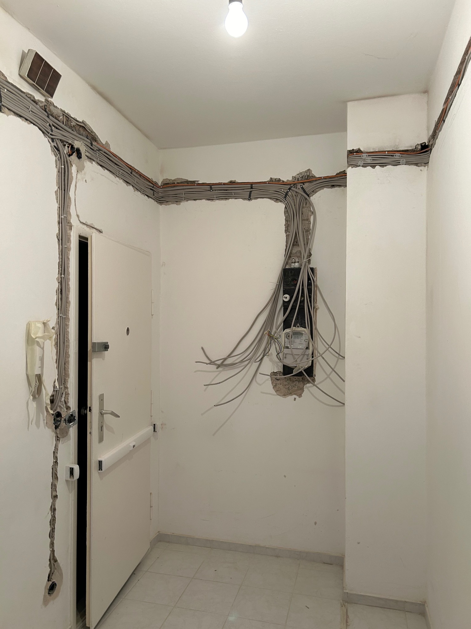 Korytarz w trakcie remontu: widoczne kable elektryczne wystające ze ściany, częściowo schowane w bruzdach, oraz skrzynka z licznikiem. Białe ściany i drzwi.