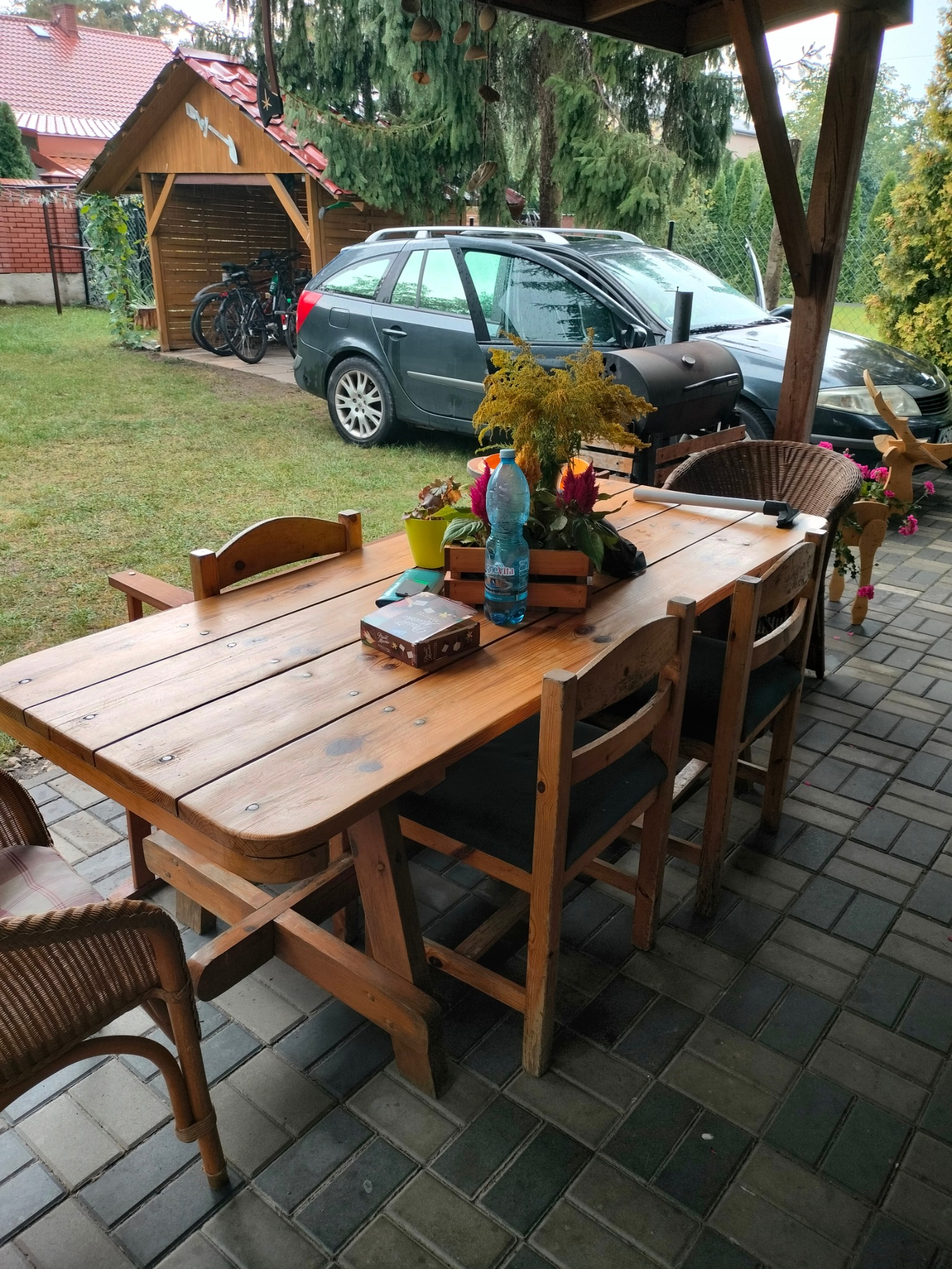 Rustykalny drewniany stół z czterema krzesłami na zewnątrz, udekorowany kwiatami, butelką wody i pudełkiem czekoladek, w tle grill, samochód i altana z rowerami.