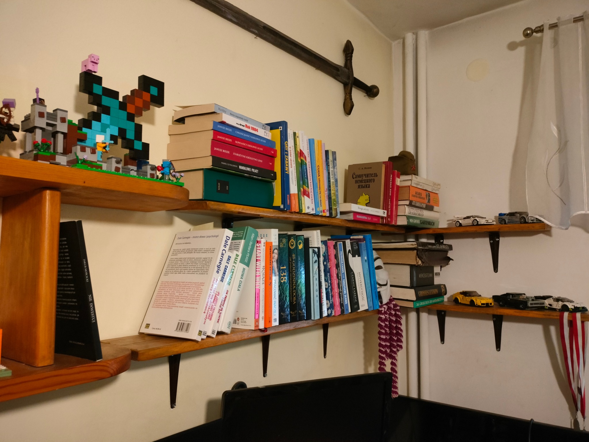 Półki z książkami, modelami samochodów i zabawkami Lego, drewniany miecz dekoracyjny na ścianie, fragment zasłony w tle.