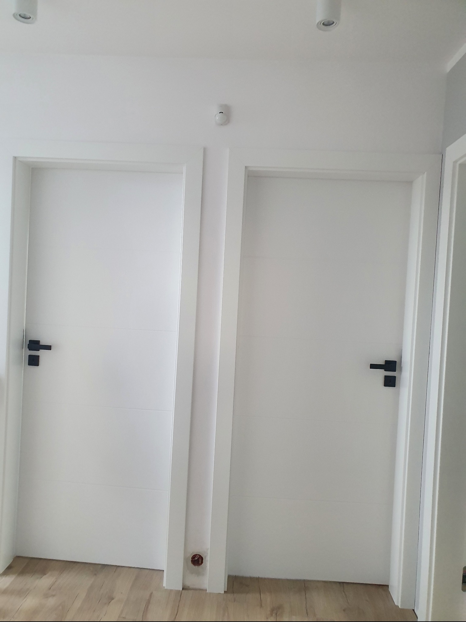 Dwa białe, minimalistyczne drzwi z czarnymi klamkami w białej futrynie, widoczne na tle jasnej ściany i podłogi imitującej drewno.