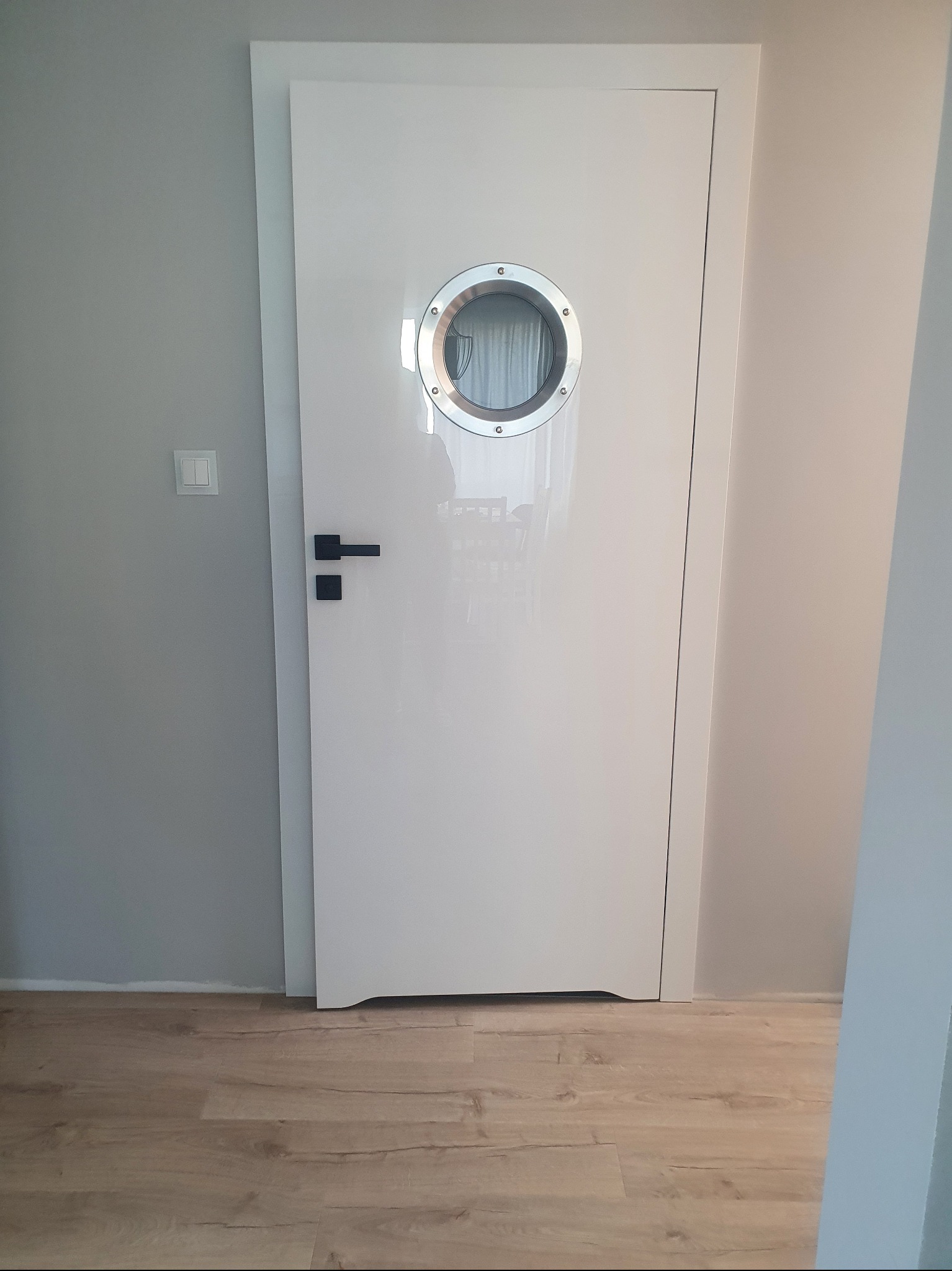 Białe, lakierowane drzwi z okrągłym, metalowym okienkiem w stylu bulaja, czarna klamka, na tle szarej ściany i paneli podłogowych.