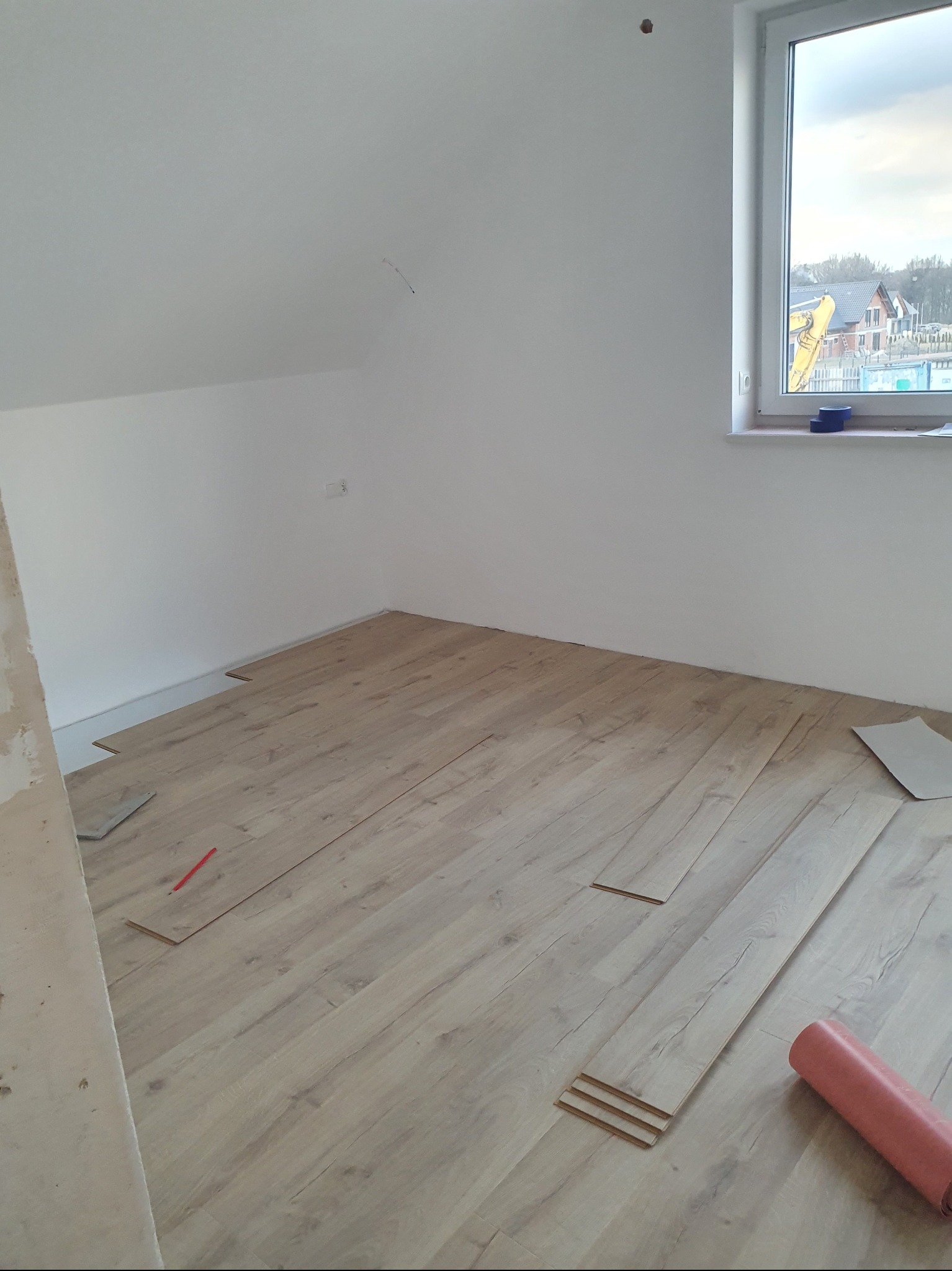 Poddasze z oknem i częściowo ułożonymi panelami podłogowymi w odcieniach beżu, widoczna rolka podkładu i narzędzia.