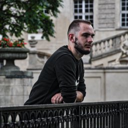 Fotograf SzeVsky - Młody mężczyzna z brodą, oparty o czarne, metalowe ogrodzenie na tle klasycystycznego budynku z rzeźbami i donicami z kwiatami.