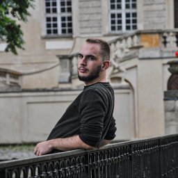 Fotograf SzeVsky - Młody mężczyzna opiera się o metalową barierkę na tle klasycystycznego budynku z donicami kwiatów, spoglądając przez ramię.