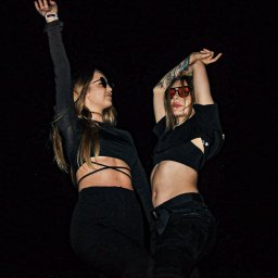 Fotograf SzeVsky - Dwie kobiety w czarnych strojach i okularach przeciwsłonecznych, z podniesionymi rękami, na ciemnym tle, podczas eventu z oświetleniem od dołu.