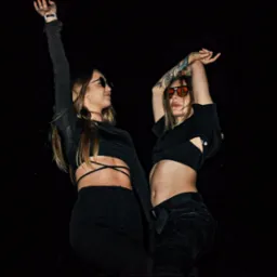 Dwie kobiety w czarnych strojach i okularach przeciwsłonecznych, z podniesionymi rękami, na ciemnym tle, podczas eventu z oświetleniem od dołu.