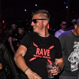 Fotograf SzeVsky - Uśmiechnięty mężczyzna w czarnych okularach i koszulce z napisem 'RAVE Dębowa Festiwal' trzymający plastikowy kubek z piwem na tle rozświetlonego tłumu w klubie.