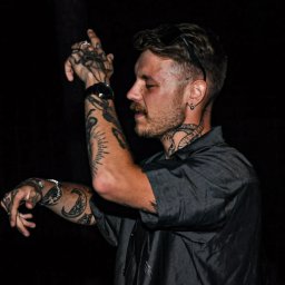 Fotograf SzeVsky - Mężczyzna z tatuażami na rękach i szyi, w szarej koszuli, z zegarkiem na lewej ręce, z okularami przeciwsłonecznymi na głowie, gestykulujący na ciemnym tle.