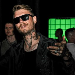 Fotograf SzeVsky - Mężczyzna z tatuażem na szyi, w czarnej koszuli i okularach przeciwsłonecznych, wskazujący palcem w Bojanowie, na tle zielonego światła i innych osób.