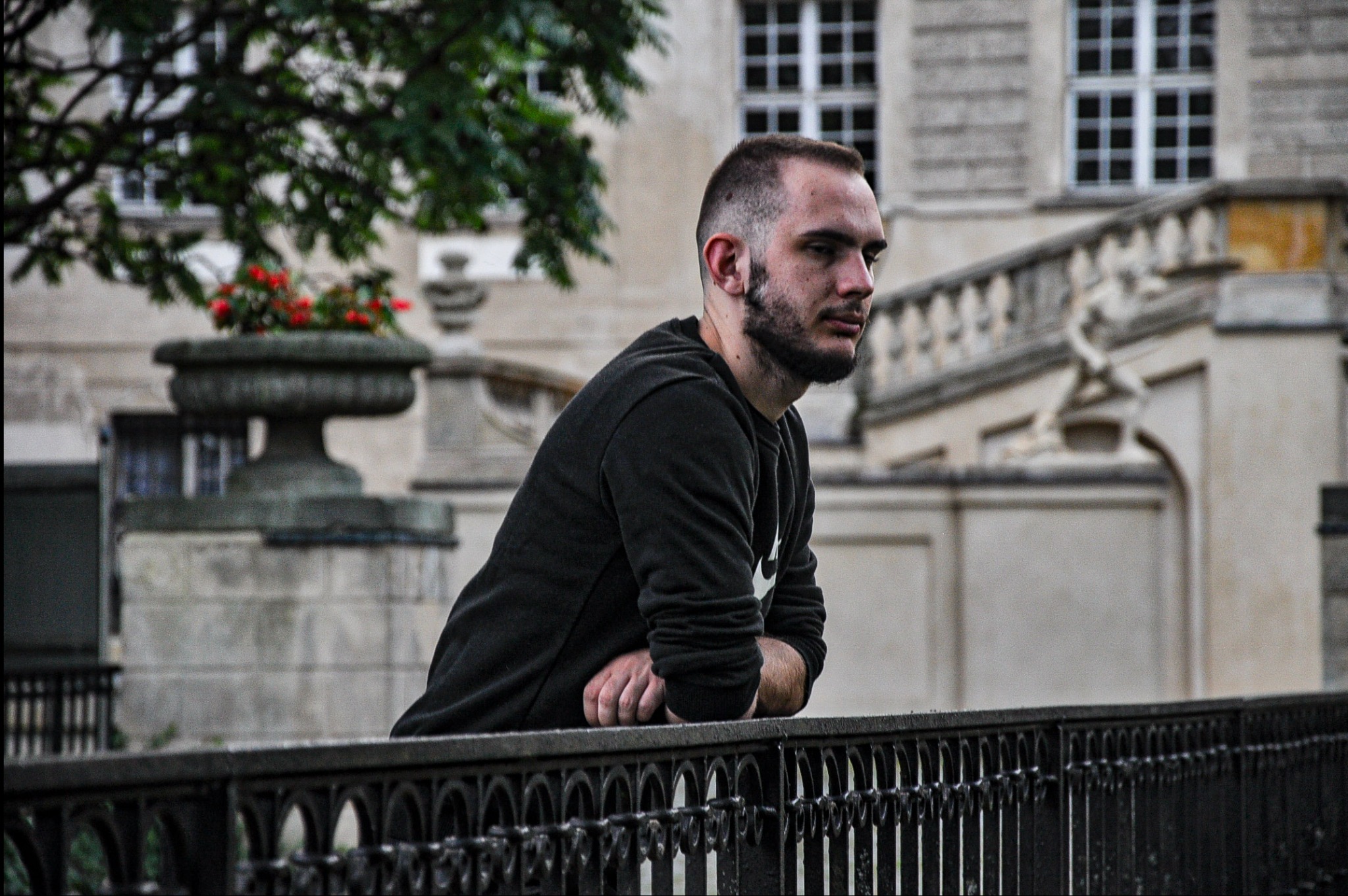 Młody mężczyzna z brodą, oparty o czarne, metalowe ogrodzenie na tle klasycystycznego budynku z rzeźbami i donicami z kwiatami.