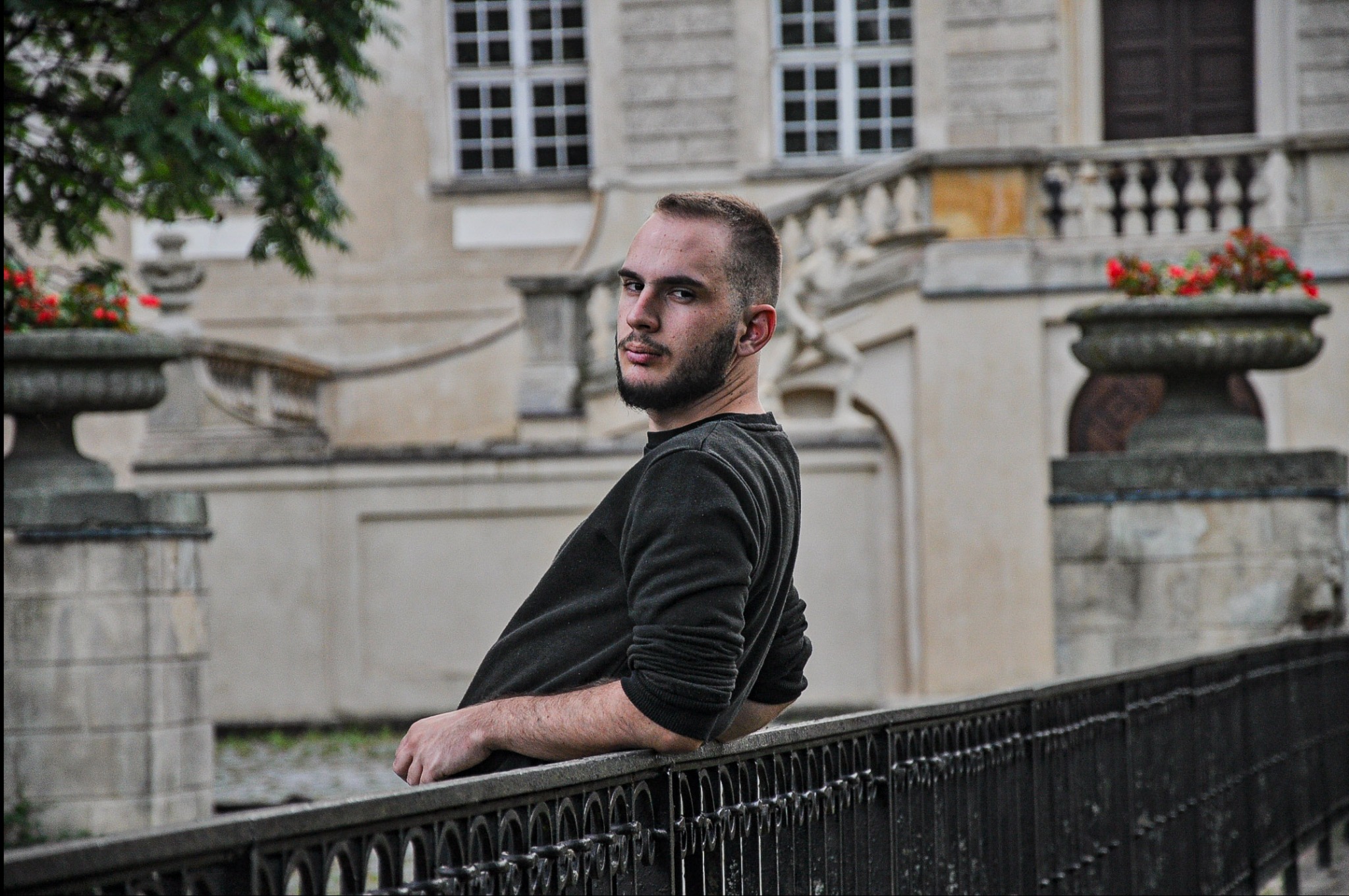 Młody mężczyzna opiera się o metalową barierkę na tle klasycystycznego budynku z donicami kwiatów, spoglądając przez ramię.