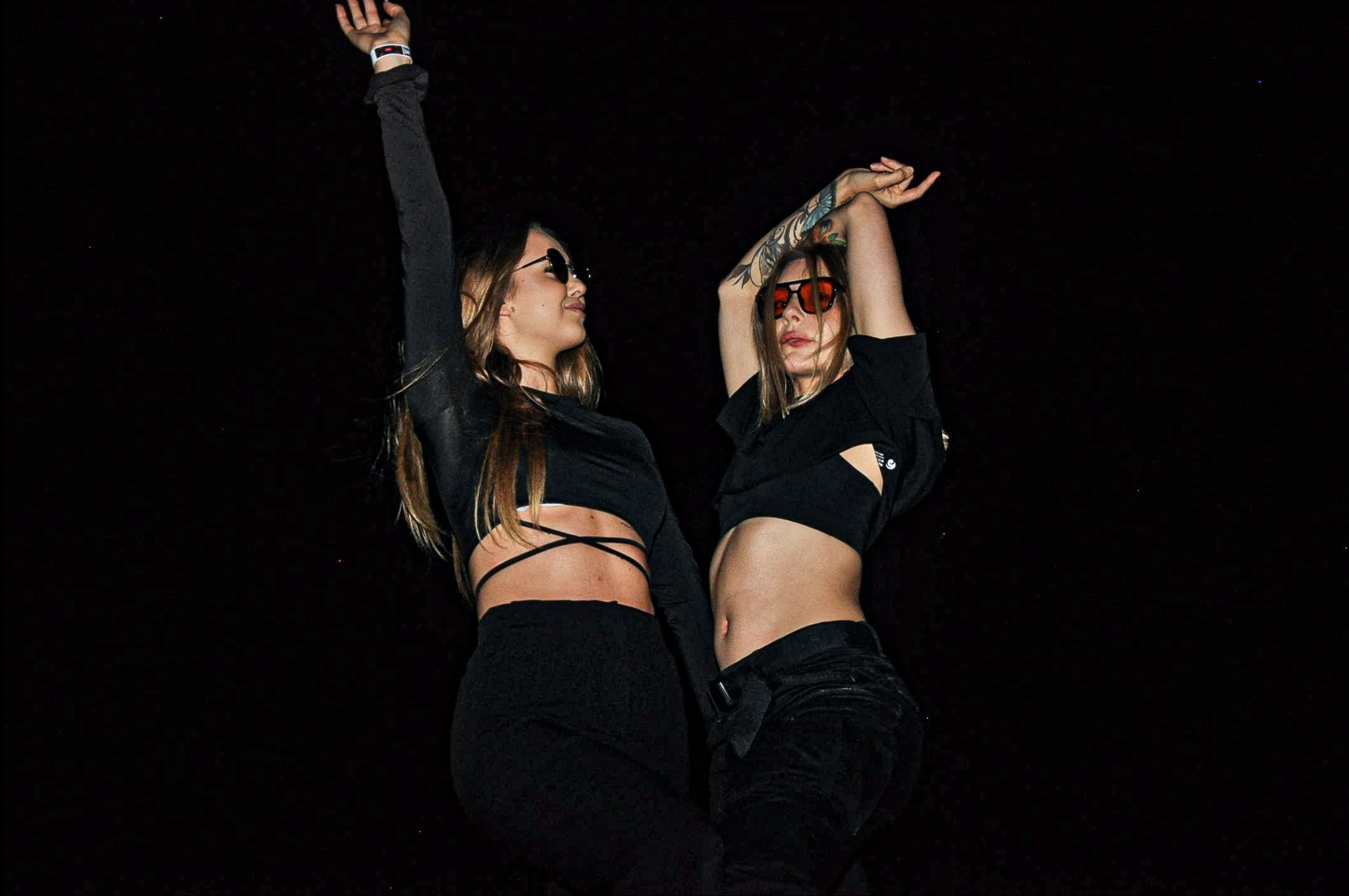Dwie kobiety w czarnych strojach i okularach przeciwsłonecznych, z podniesionymi rękami, na ciemnym tle, podczas eventu z oświetleniem od dołu.