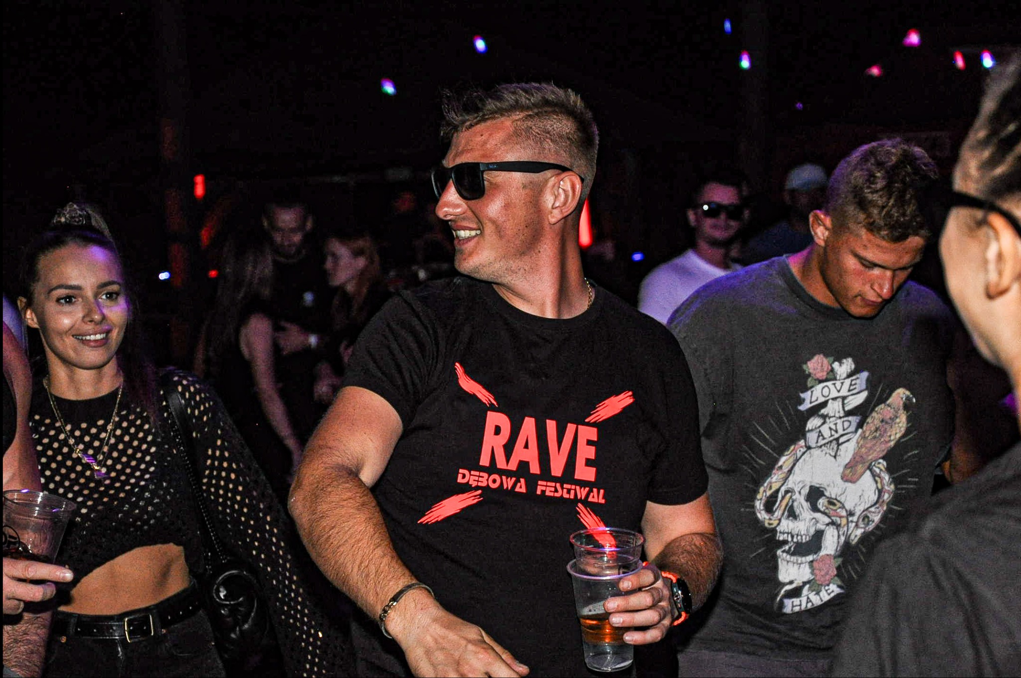 Uśmiechnięty mężczyzna w czarnych okularach i koszulce z napisem 'RAVE Dębowa Festiwal' trzymający plastikowy kubek z piwem na tle rozświetlonego tłumu w klubie.
