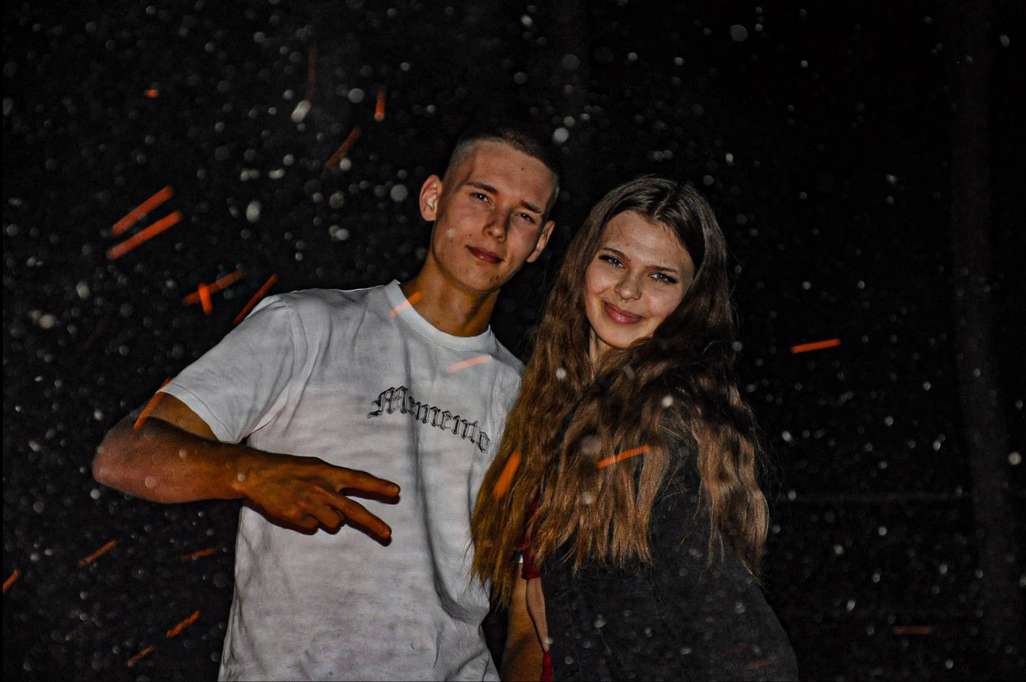 Młoda para pozuje na tle ciemnego nieba z efektem bokeh, mężczyzna w białej koszulce z napisem 'Memento' pokazuje znak pokoju.