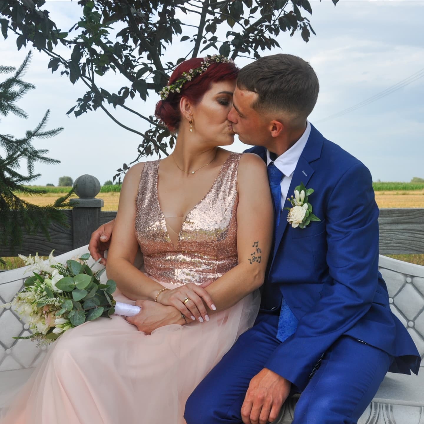 Para młoda całuje się siedząc na ławce; panna młoda w sukni z cekinami i wiankiem, pan młody w niebieskim garniturze, bukiet ślubny na ławce, w tle pole.