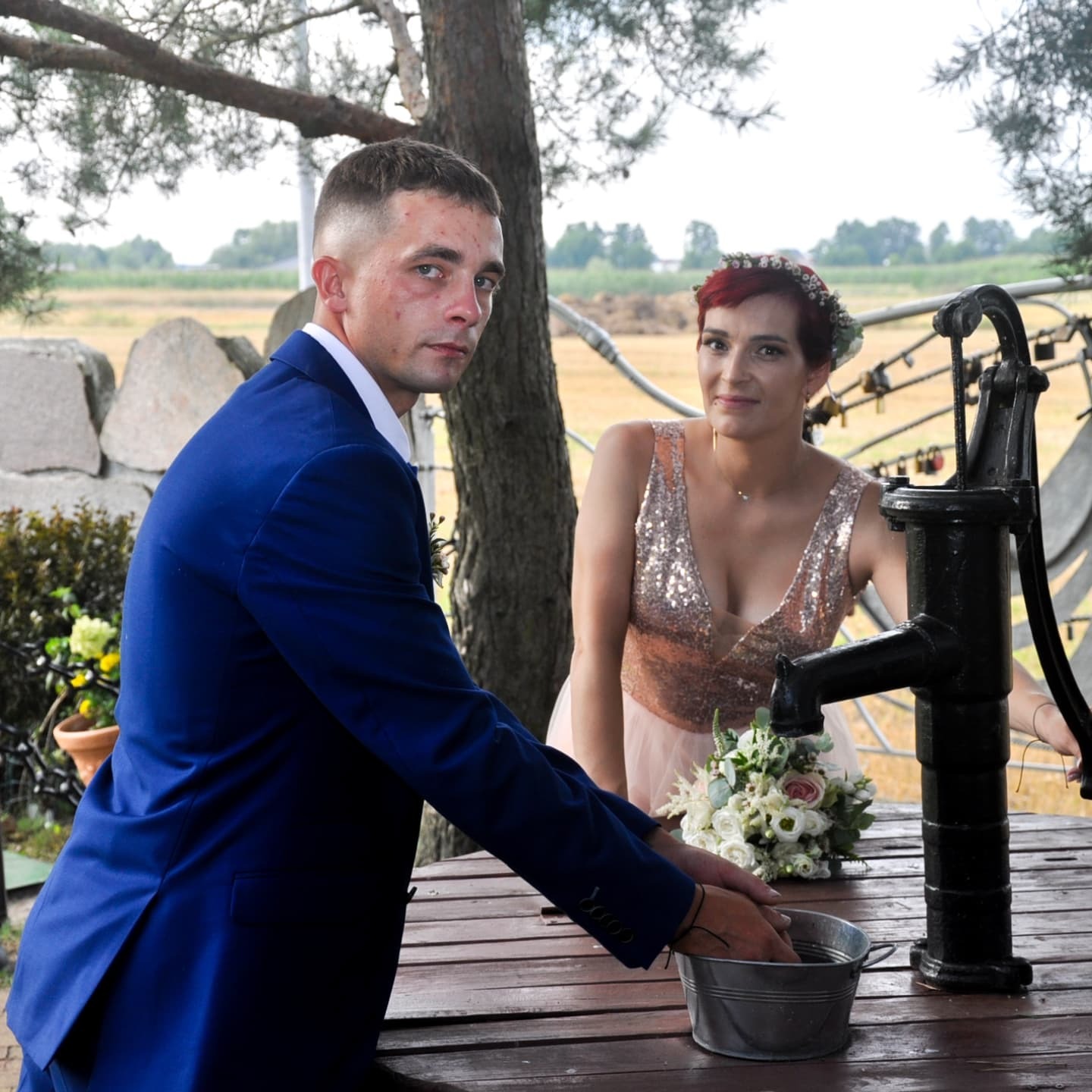 Para młoda przy studni: pan młody w granatowym garniturze myje ręce w wiadrze, panna młoda w sukni z cekinami i wiankiem na głowie, bukiet ślubny na stole.