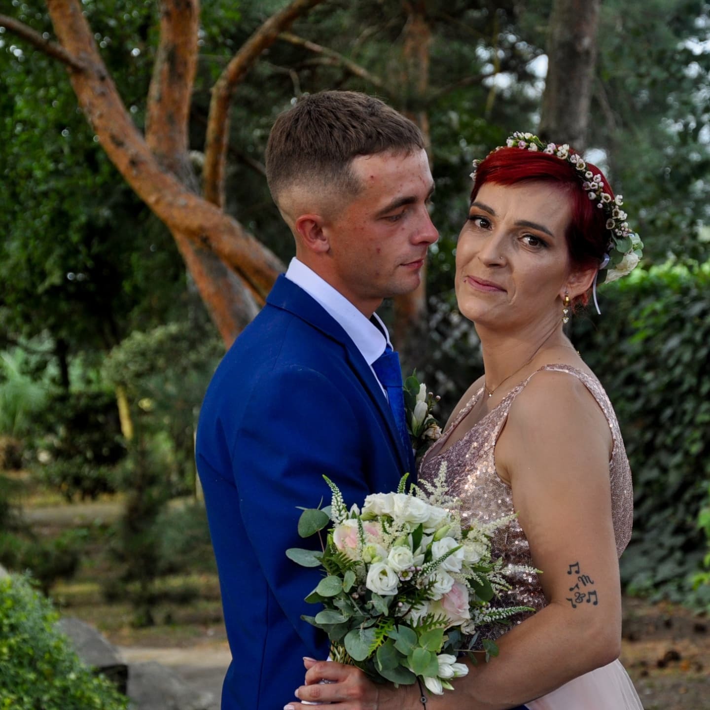 Para młoda: mężczyzna w niebieskim garniturze spogląda na kobietę w sukience z cekinami i wiankiem, trzymającą bukiet, widoczne tatuaże na ramieniu kobiety.