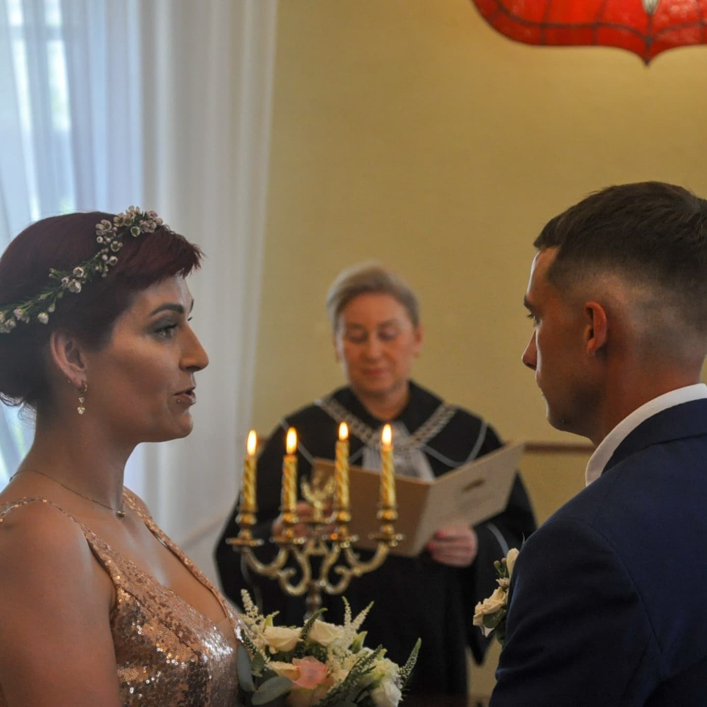 Panna młoda w sukni z cekinami i wiankiem na głowie podczas ceremonii ślubnej, obok stoi pan młody, w tle urzędniczka czyta tekst z księgi, a przed nią płoną świece.