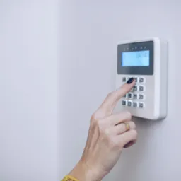 Kobieta z pierścionkiem na palcu wprowadza kod do panelu alarmowego z podświetlanym wyświetlaczem LCD, zamontowanego na białej ścianie.