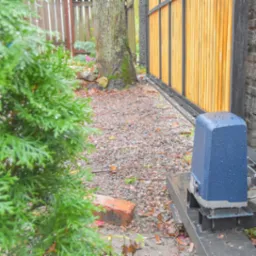 Niebieski napęd do bramy przesuwnej zamontowany na metalowej podstawie, widoczne krople deszczu, brama z jasnego drewna i czarnych profili, tło z drzewem i krzewami.