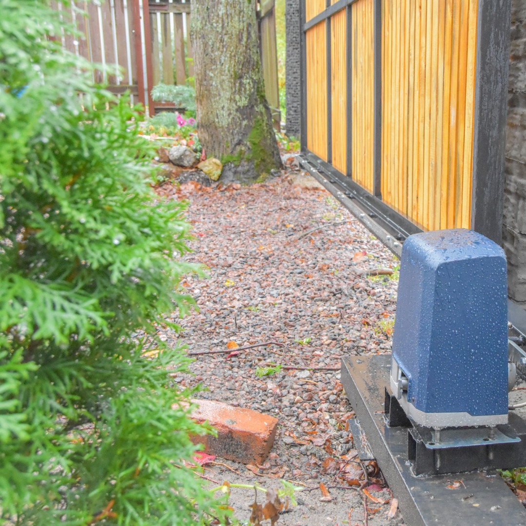 Niebieski napęd do bramy przesuwnej zamontowany na metalowej podstawie, widoczne krople deszczu, brama z jasnego drewna i czarnych profili, tło z drzewem i krzewami.