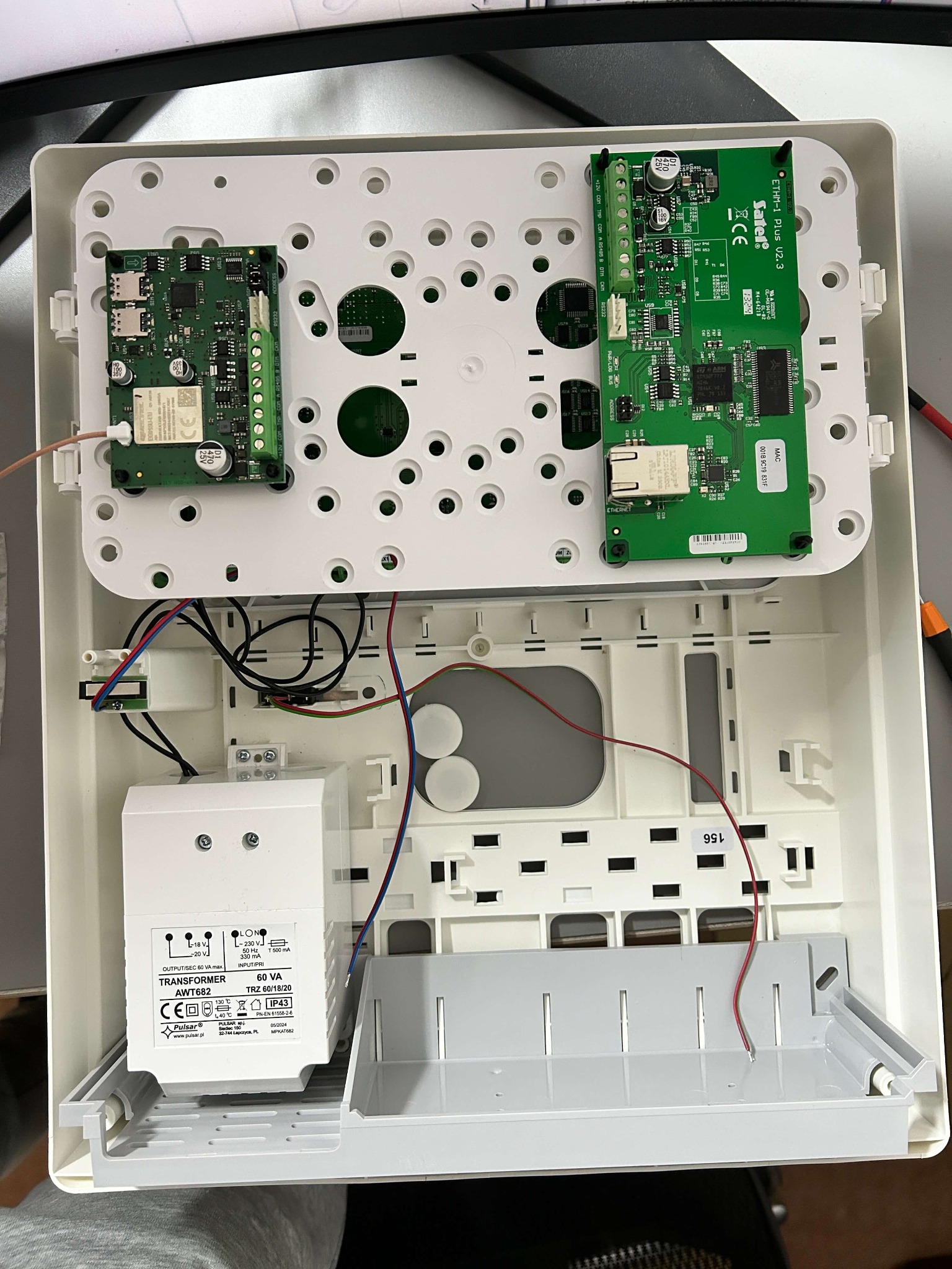 Otwarta obudowa centrali alarmowej z widocznymi modułami elektronicznymi, transformatorem i okablowaniem w trakcie montażu.