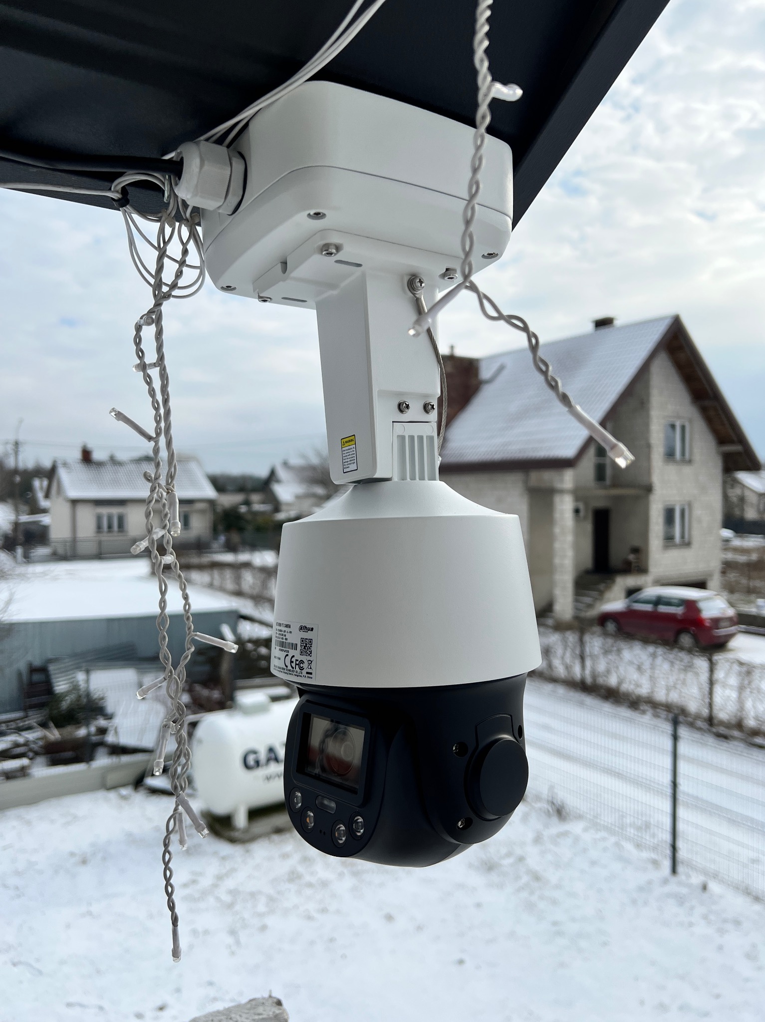 Zewnętrzna kamera obrotowa PTZ zamontowana na zewnątrz budynku, widoczne okablowanie i dekoracja w postaci łańcucha świetlnego, w tle ośnieżony krajobraz z domami i zbiornikiem gazu.