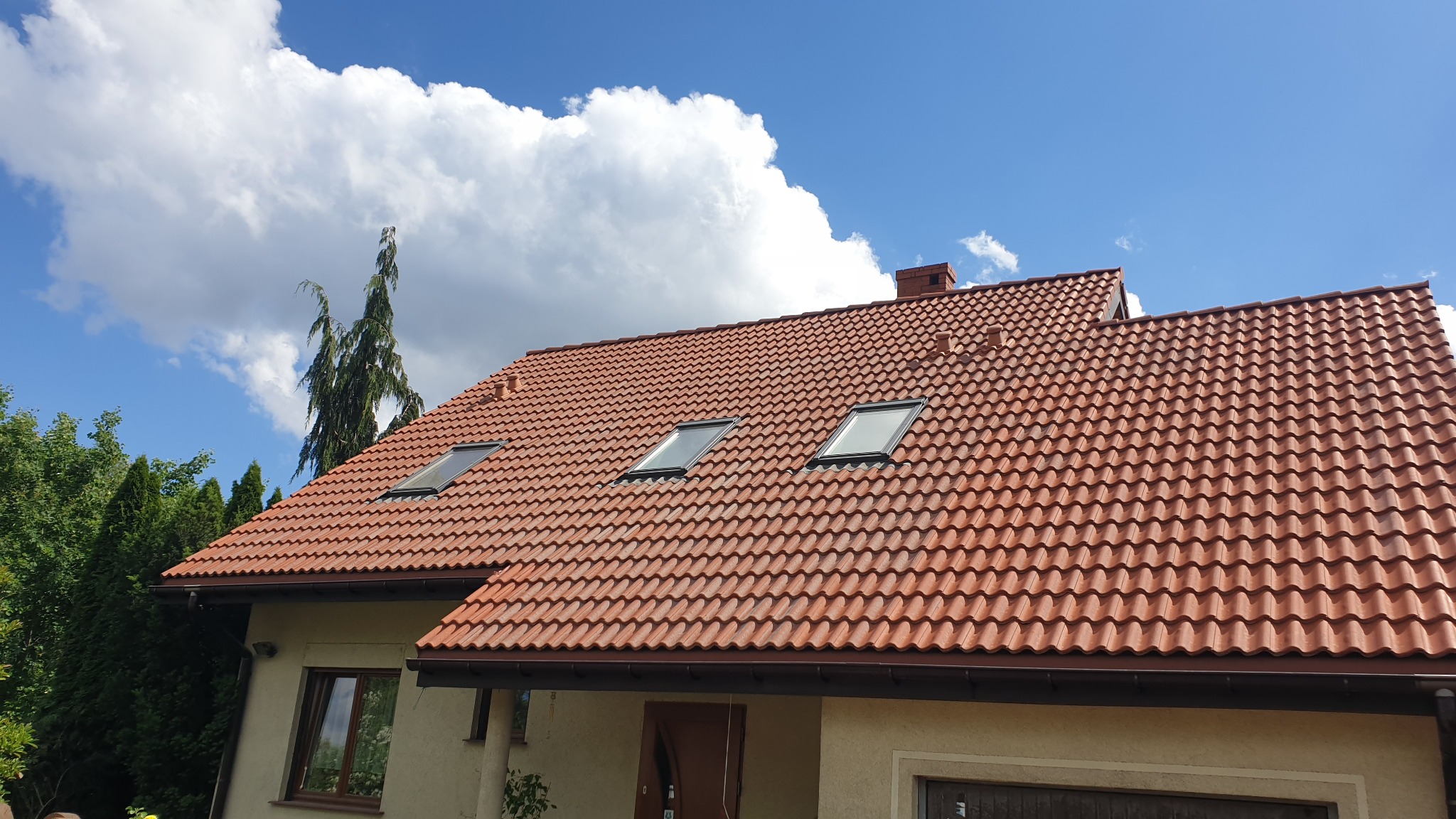 Czerwony dach z dachówki ceramicznej z trzema oknami dachowymi, widok z dołu na tle błękitnego nieba z białymi chmurami i zieleni drzew.