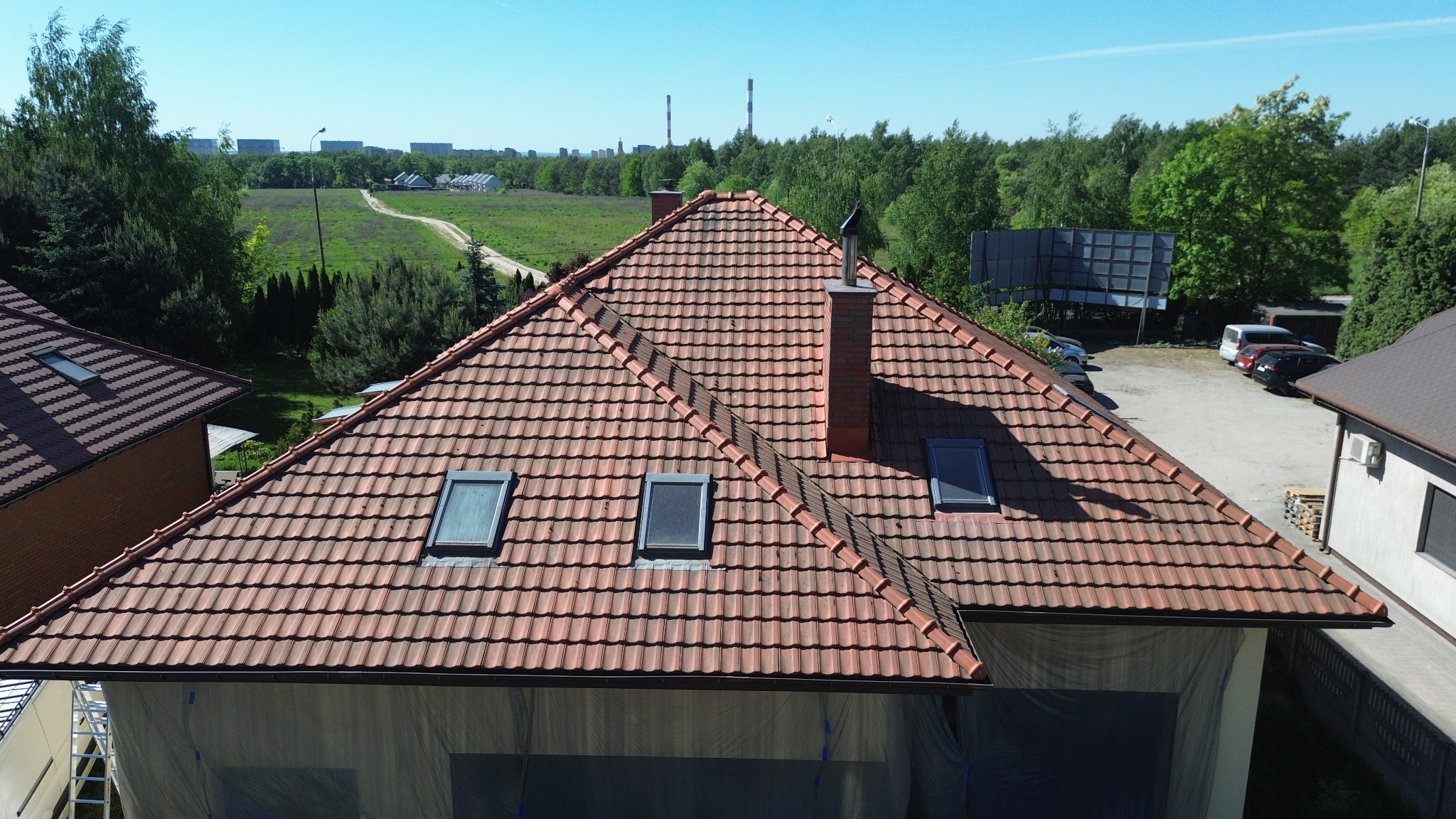 Czerwona dachówka ceramiczna na dachu domu jednorodzinnego z trzema oknami dachowymi i kominem z cegły, widok z góry, w tle zieleń i kominy przemysłowe.