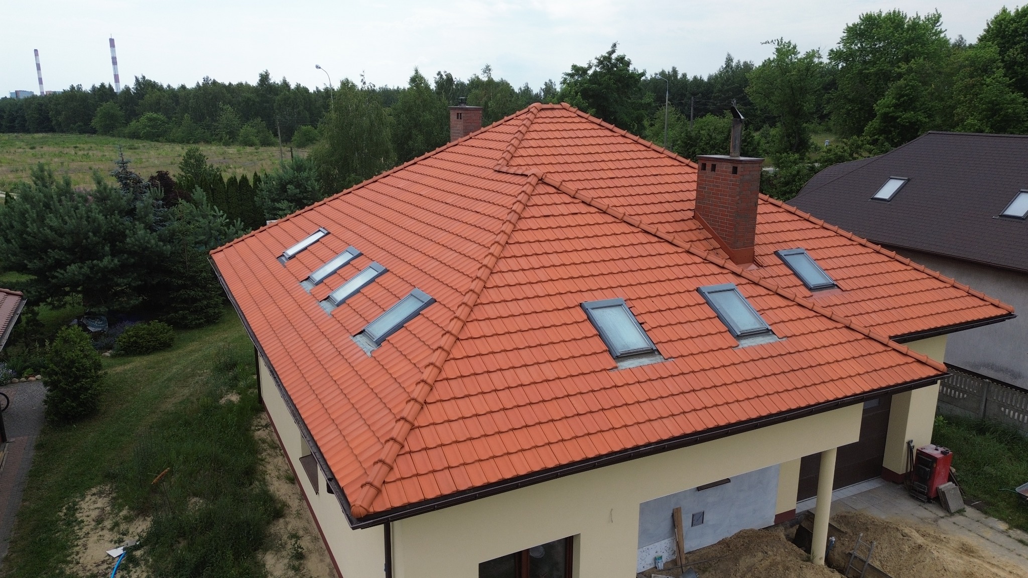 Widok z góry na dom z nową, czerwoną dachówką ceramiczną i oknami dachowymi; w tle widoczne drzewa i komin elektrowni.