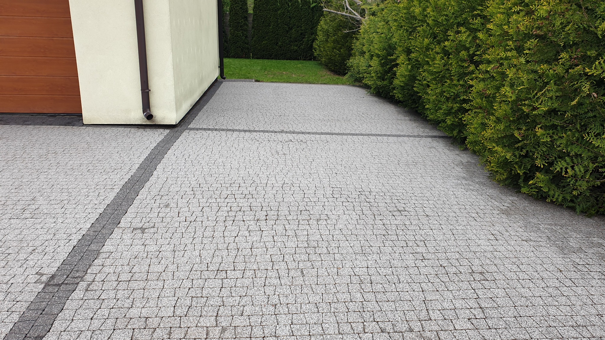 Szara kostka brukowa ułożona w jodełkę, obrzeża z ciemniejszej kostki, budynek z brązową bramą garażową i rynną, żywopłot z tui w tle.