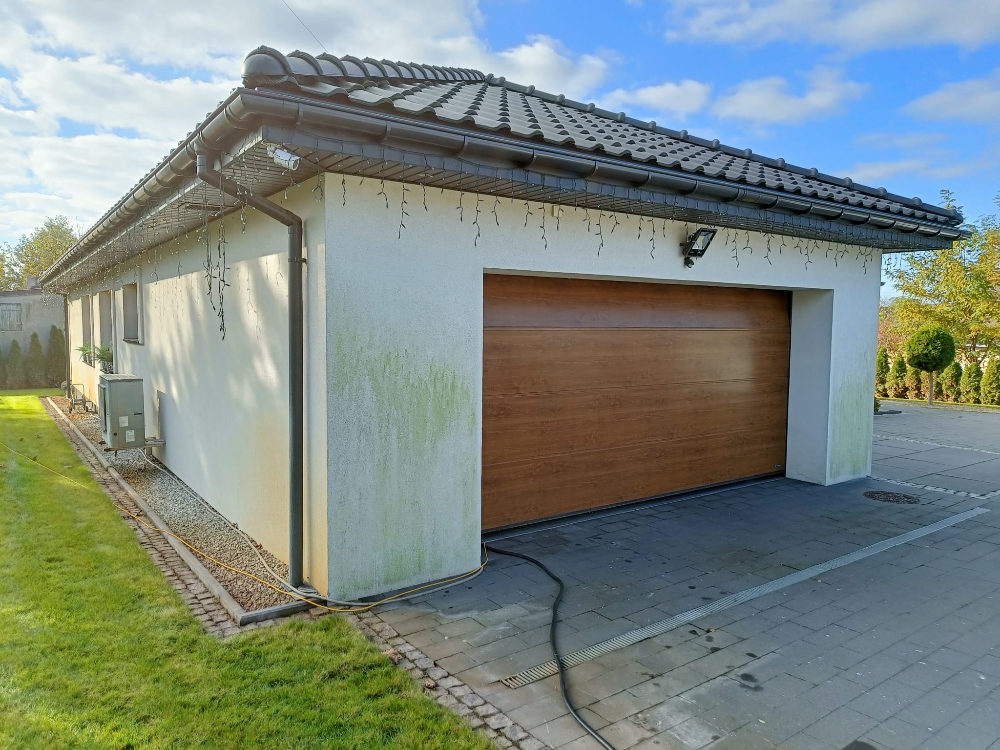 Boczny widok garażu z brązową bramą segmentową, szarą dachówką i ciemnymi rynnami, elewacja z zielonym nalotem, na kostce brukowej leżą kable.