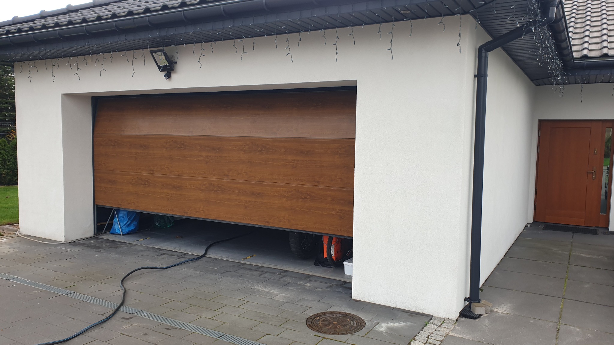 Nowoczesny garaż z częściowo otwartą, segmentową bramą w kolorze drewna, z zewnętrznym oświetleniem i dekoracyjnymi lampkami na dachu, widoczne elementy elewacji i rynny.