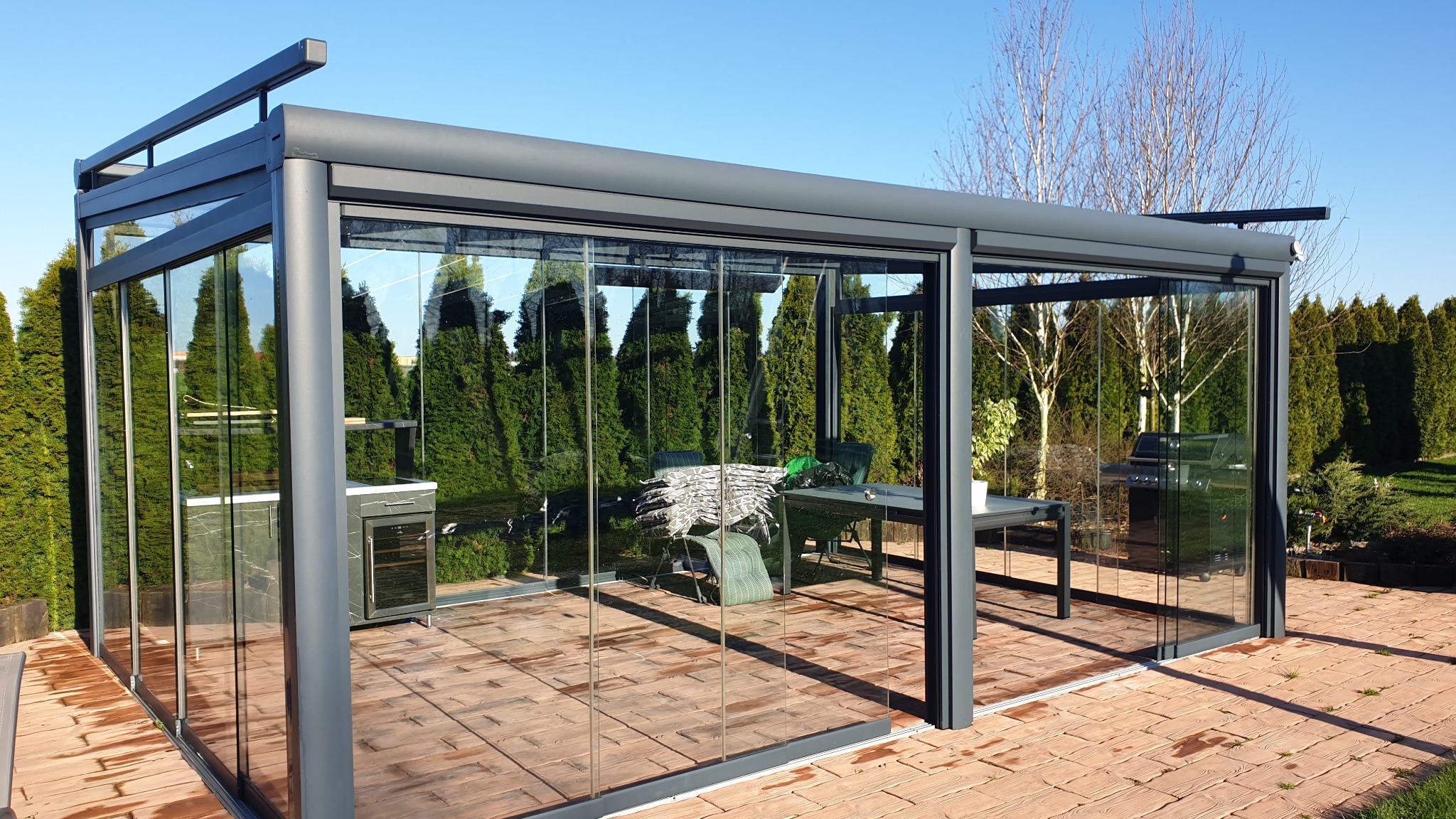 Nowoczesna szklana altana z przesuwnymi drzwiami, wyposażona w meble ogrodowe i grill, otoczona zielonymi tujami i brukowanym tarasem.