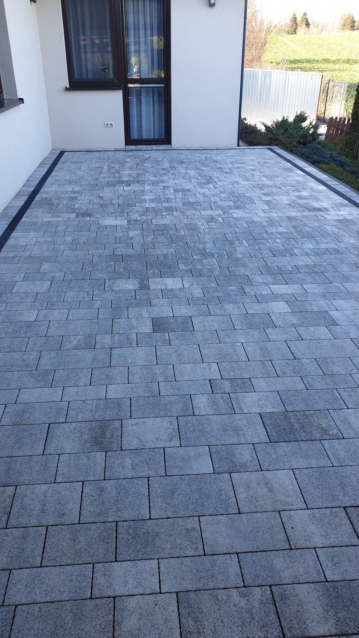 Świeżo ułożona, szara kostka brukowa w regularnym wzorze, przylegająca do białej ściany budynku z oknem. Czarny pas oddziela kostkę od ściany. W tle widoczny fragment ogrodu i metalowe ogrodzenie.