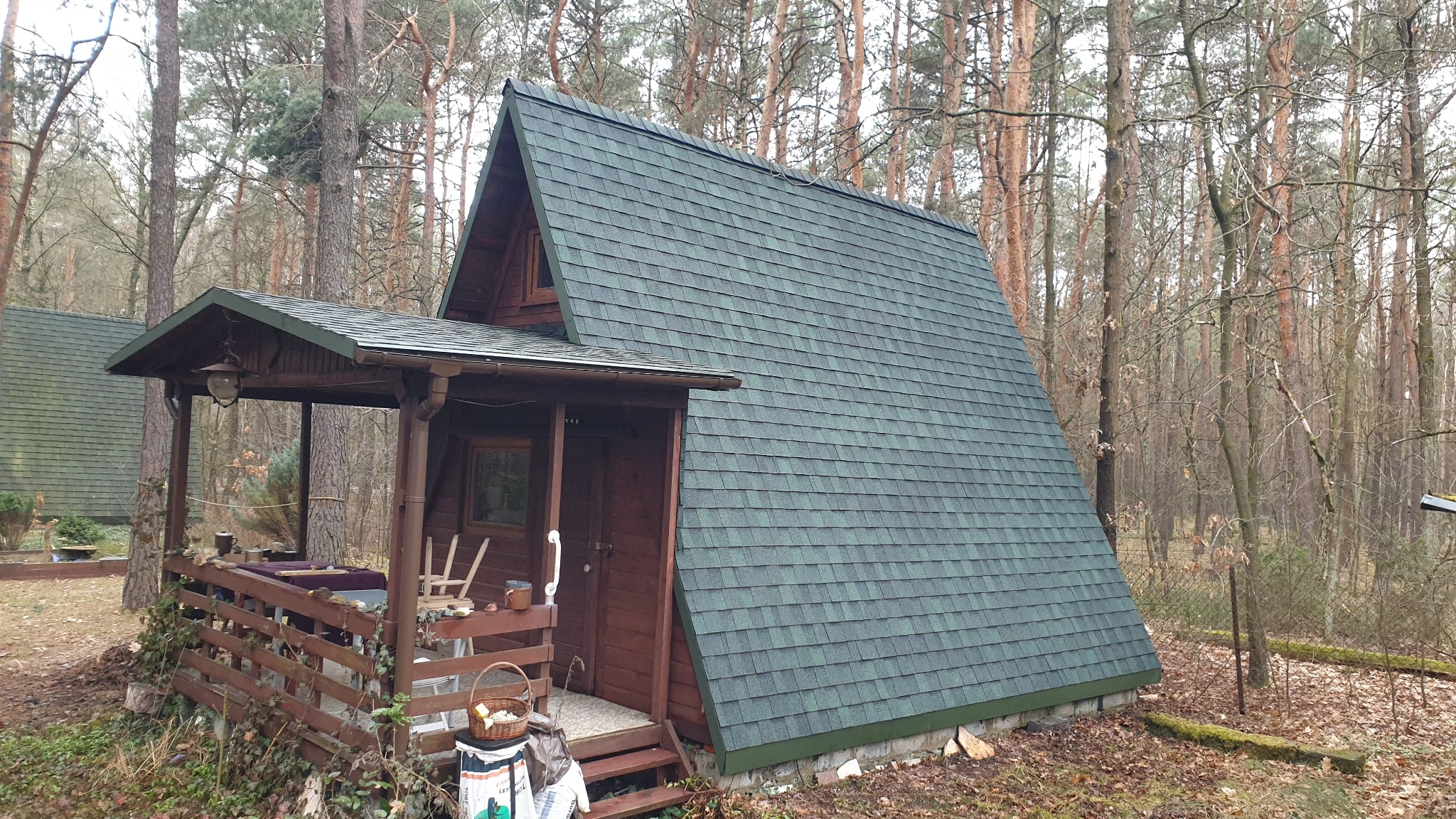 Domek letniskowy typu A-frame z nowym, zielonym dachem z gontu bitumicznego, otoczony sosnowym lasem w pochmurny dzień.