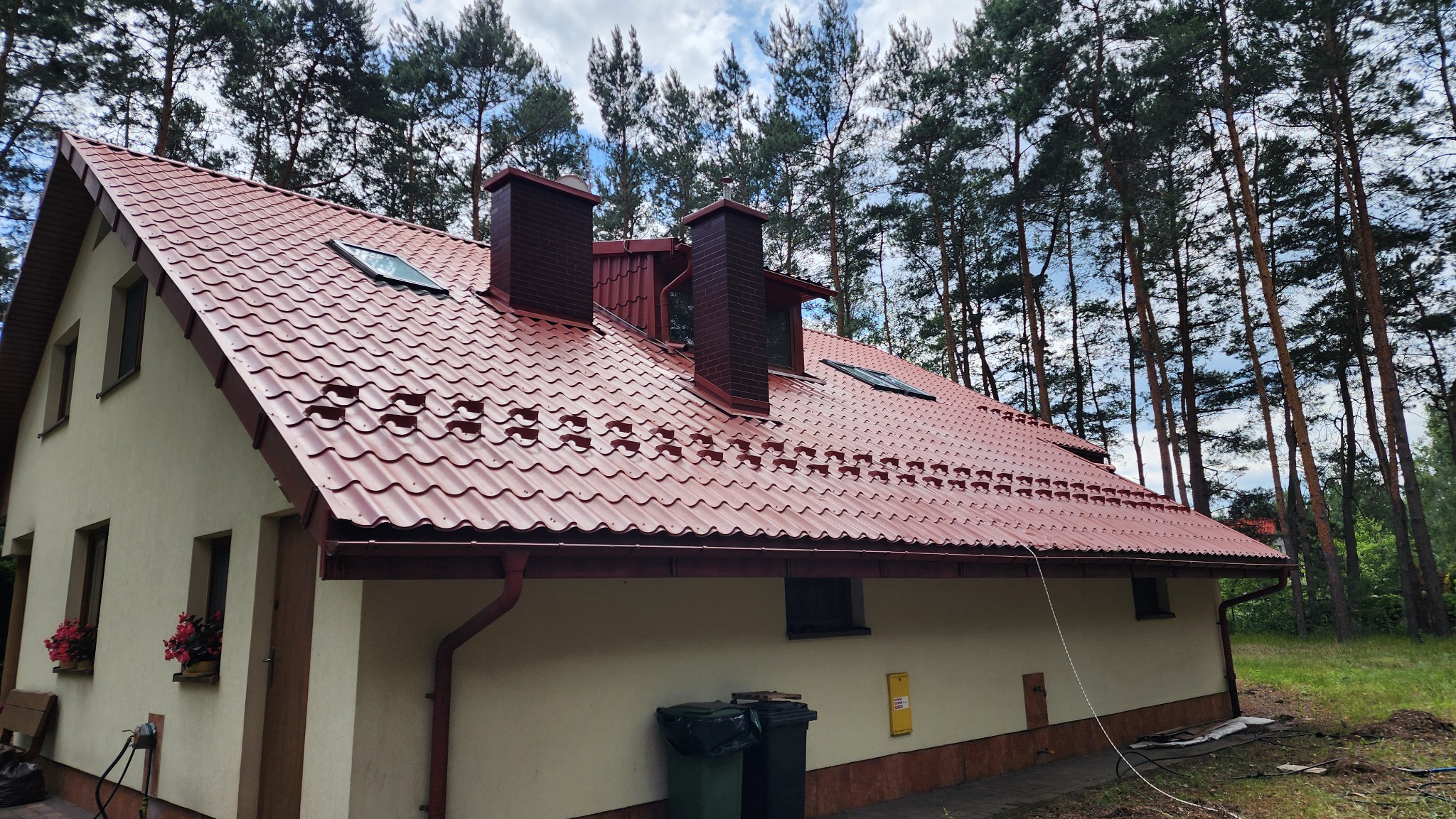 Dom jednorodzinny z nowym, czerwonym dachem z blachodachówki, widoczne kominy z cegły i rynny. W tle wysokie drzewa iglaste.