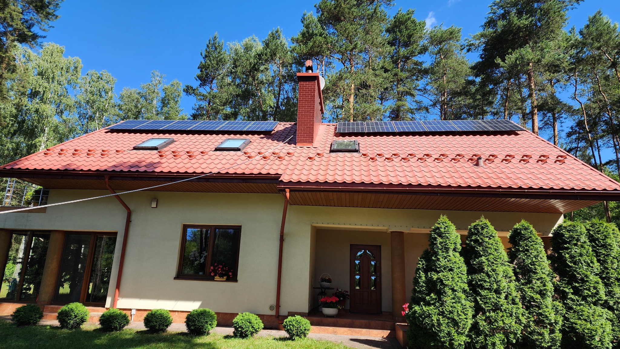 Dom z czerwoną dachówką, panelami solarnymi i ceglanym kominem na tle zielonych drzew i błękitnego nieba. Widoczne okna dachowe i zadbany ogród.