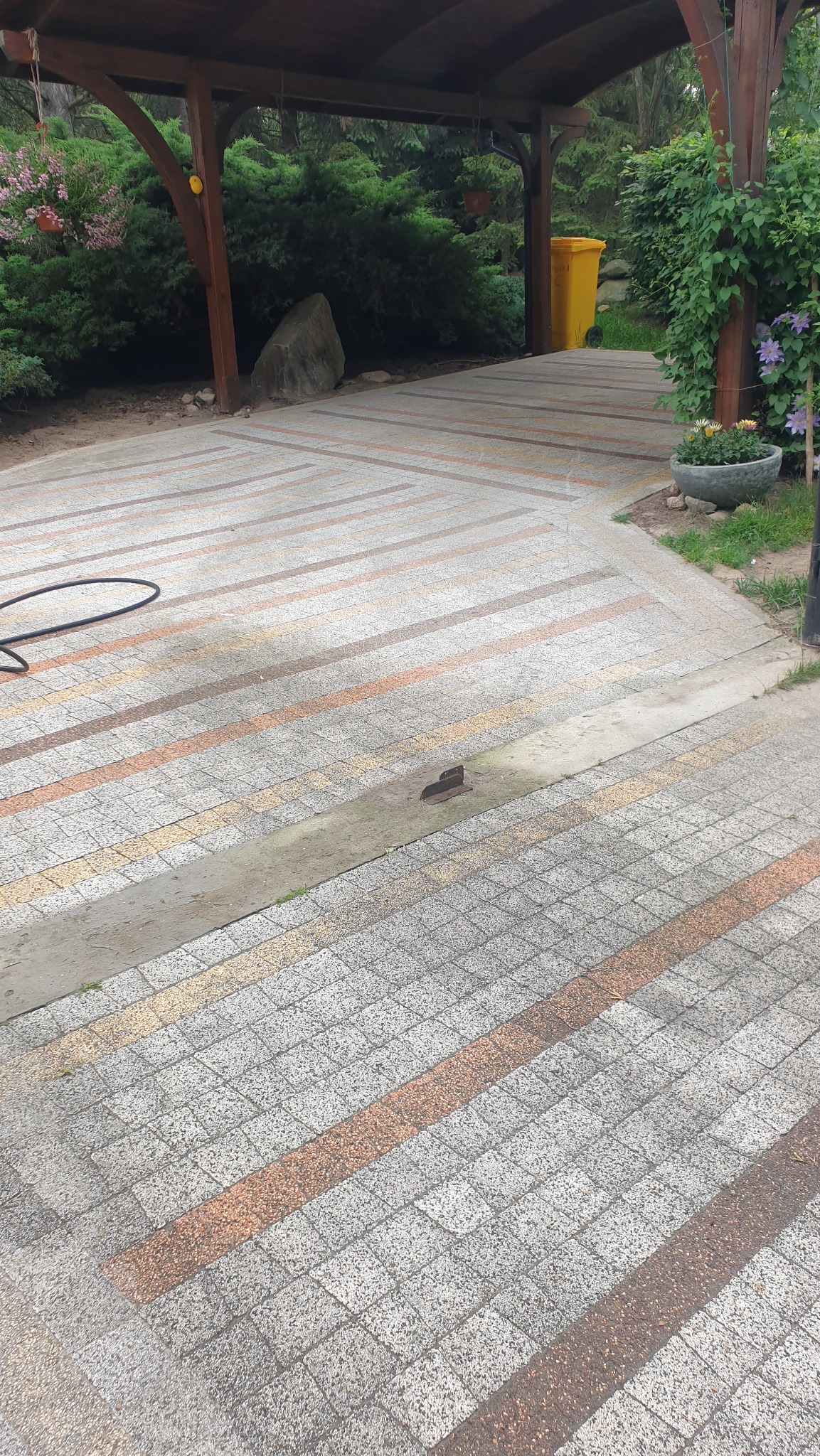 Kostka brukowa ułożona w pasy pod drewnianą konstrukcją. Widoczny wąż ogrodowy i żółty kosz na śmieci w tle. Część kostki jest mokra, co sugeruje niedawne mycie.