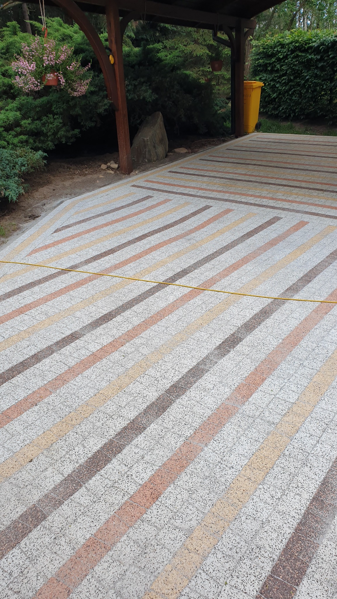 Wzorzysta kostka brukowa na podjeździe, ułożona w pasy z wykorzystaniem różnych odcieni. W tle widoczny fragment zadaszenia i roślinność.