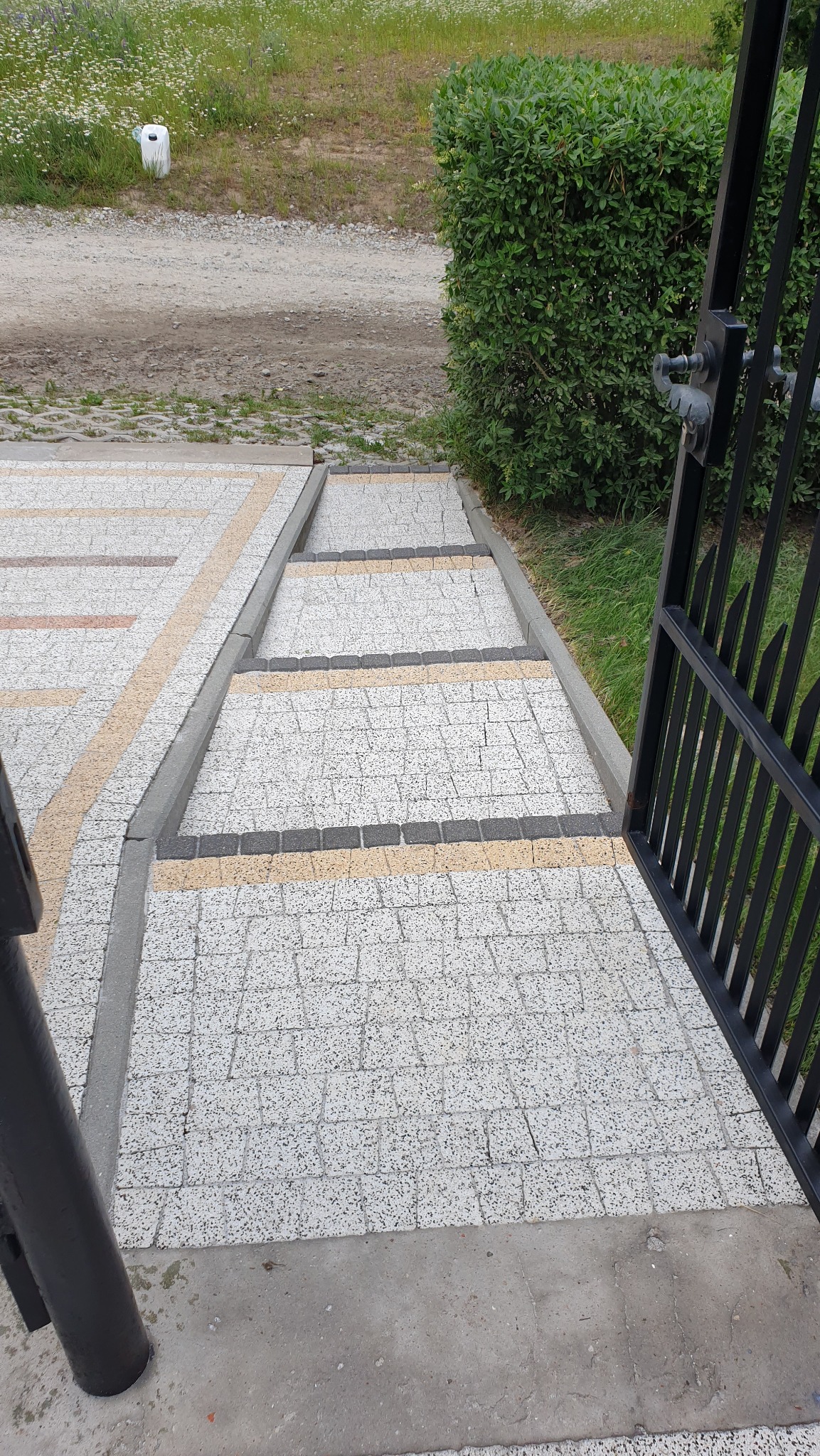 Schody z kostki brukowej w odcieniach szarości i beżu, prowadzące do ogrodu z otwartą, czarną bramą. Widok z góry.