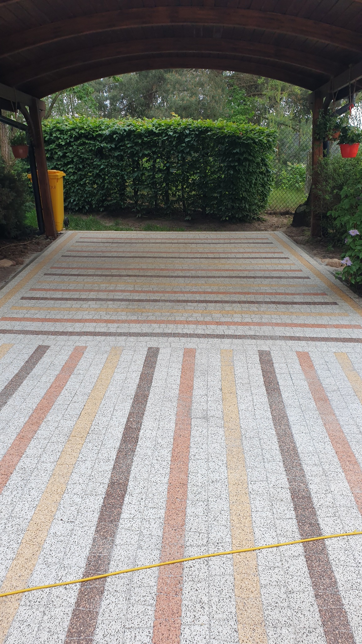 Wzorzysta kostka brukowa pod zadaszeniem, z żółtym wężem ogrodowym i zielonym żywopłotem w tle. Widok z perspektywy.