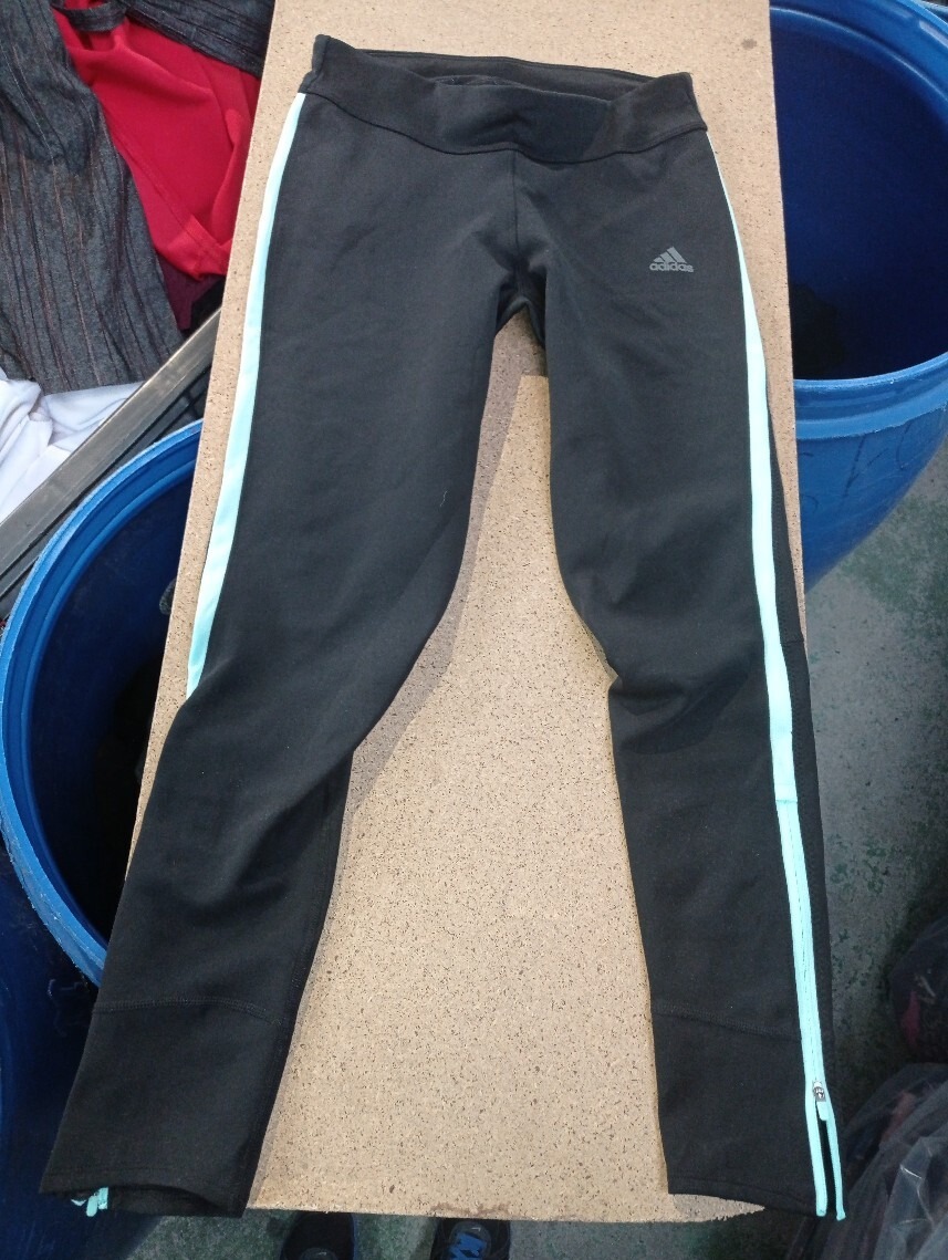 Czarne legginsy sportowe Adidas z jasnoniebieskim paskiem bocznym i zamkiem błyskawicznym u dołu, leżące na tekturowej powierzchni, z fragmentami niebieskiego wiadra i ubrań w tle.