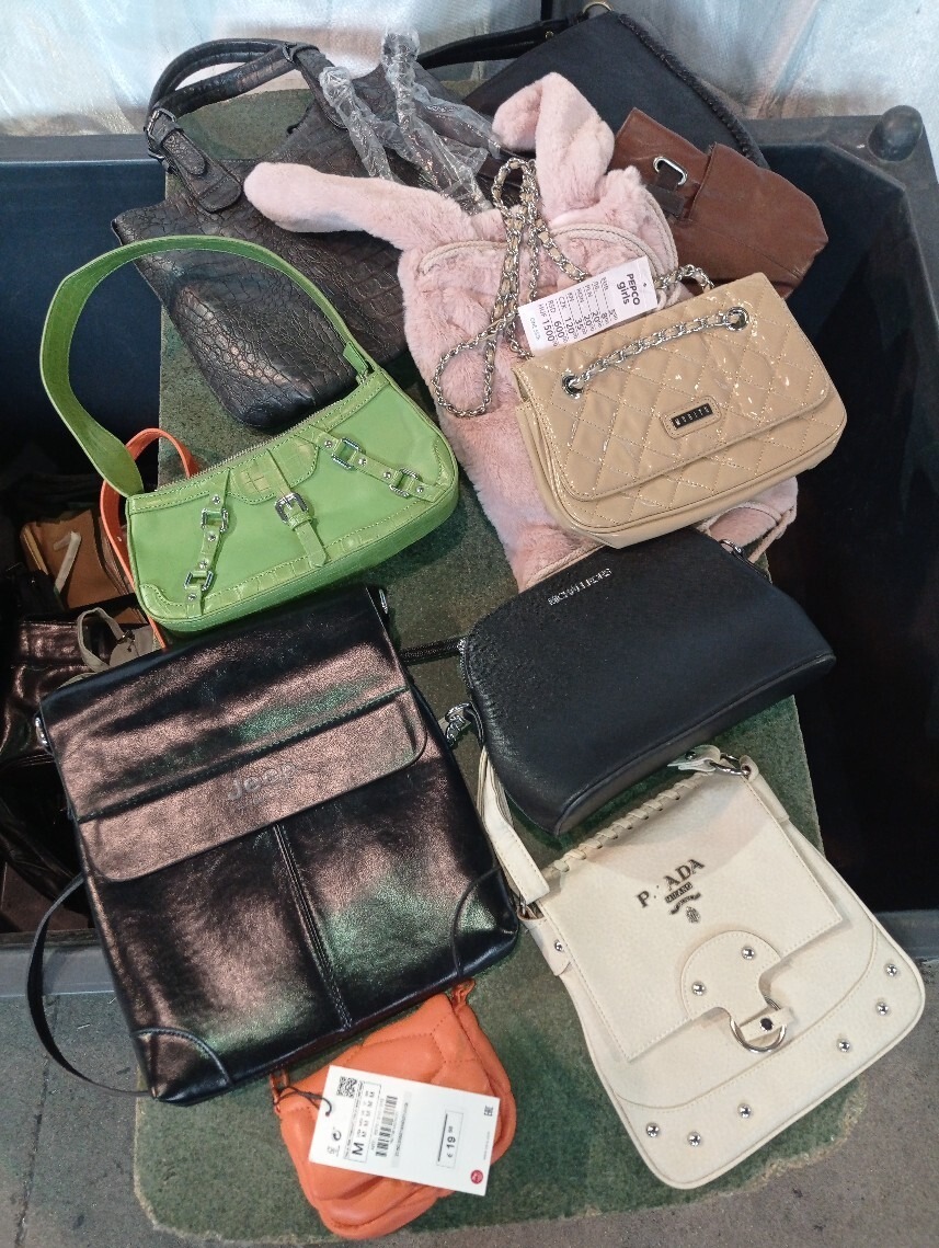 Różnorodny zbiór torebek używanych, w tym zielona z ozdobnymi klamrami, beżowa pikowana na łańcuszku, czarna skórzana typu listonoszka i kremowa z logo Prada, ułożonych na podłodze.
